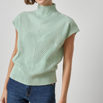 A woman wearing a light sage green ribbed knit mock neck top with cap sleeves and blue jeans, photographed against a neutral light gray background.