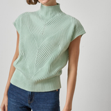 A woman wearing a light sage green ribbed knit mock neck top with cap sleeves and blue jeans, photographed against a neutral light gray background.