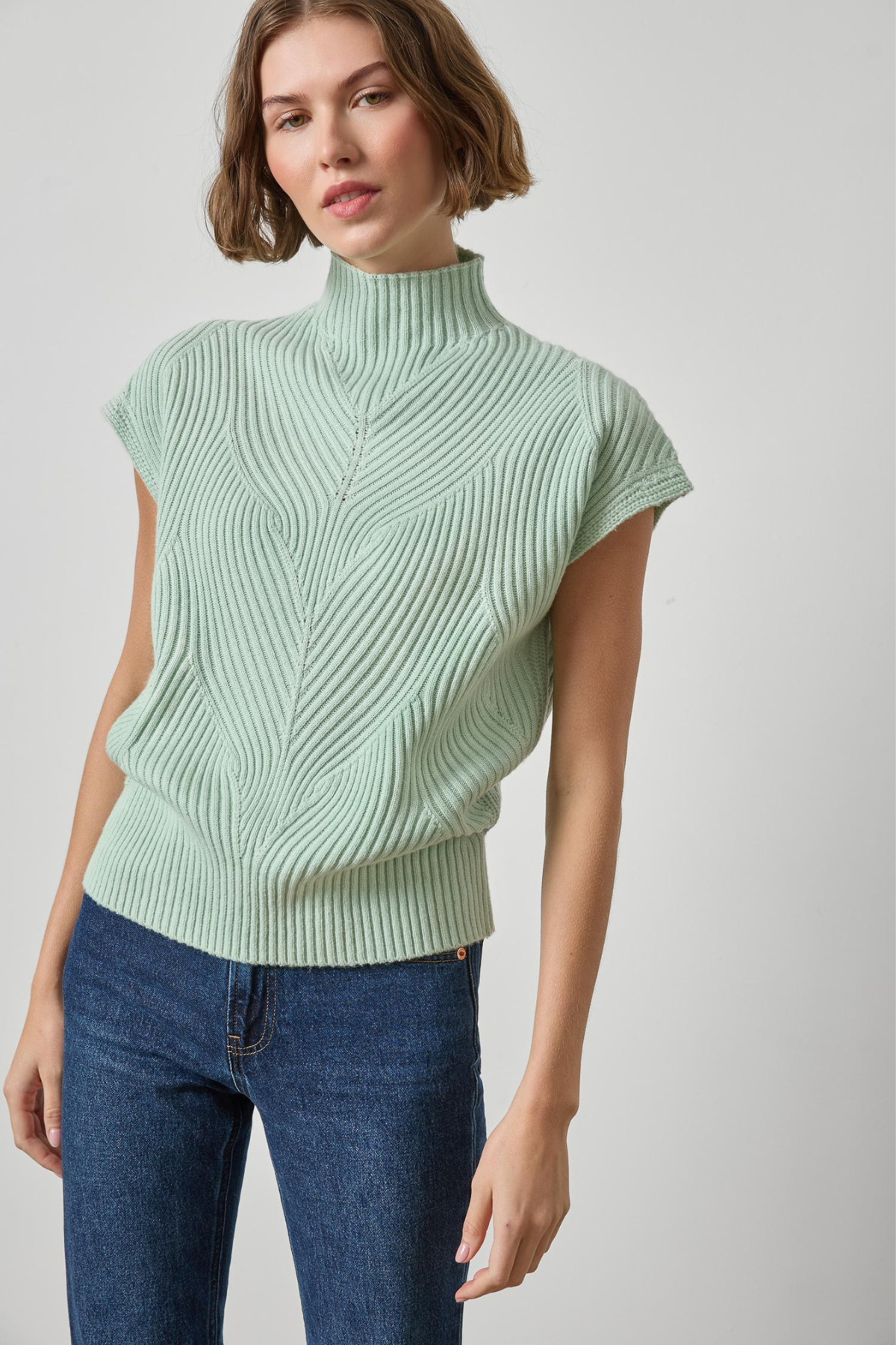 A woman wearing a light sage green ribbed knit mock neck top with cap sleeves and blue jeans, photographed against a neutral light gray background.