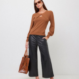 A woman modeling a brown long-sleeve knit top with a crossover front and keyhole cutout, paired with black wide-leg pants, leopard-print heeled mules, sunglasses, and a brown textured tote bag against a plain light gray studio background.