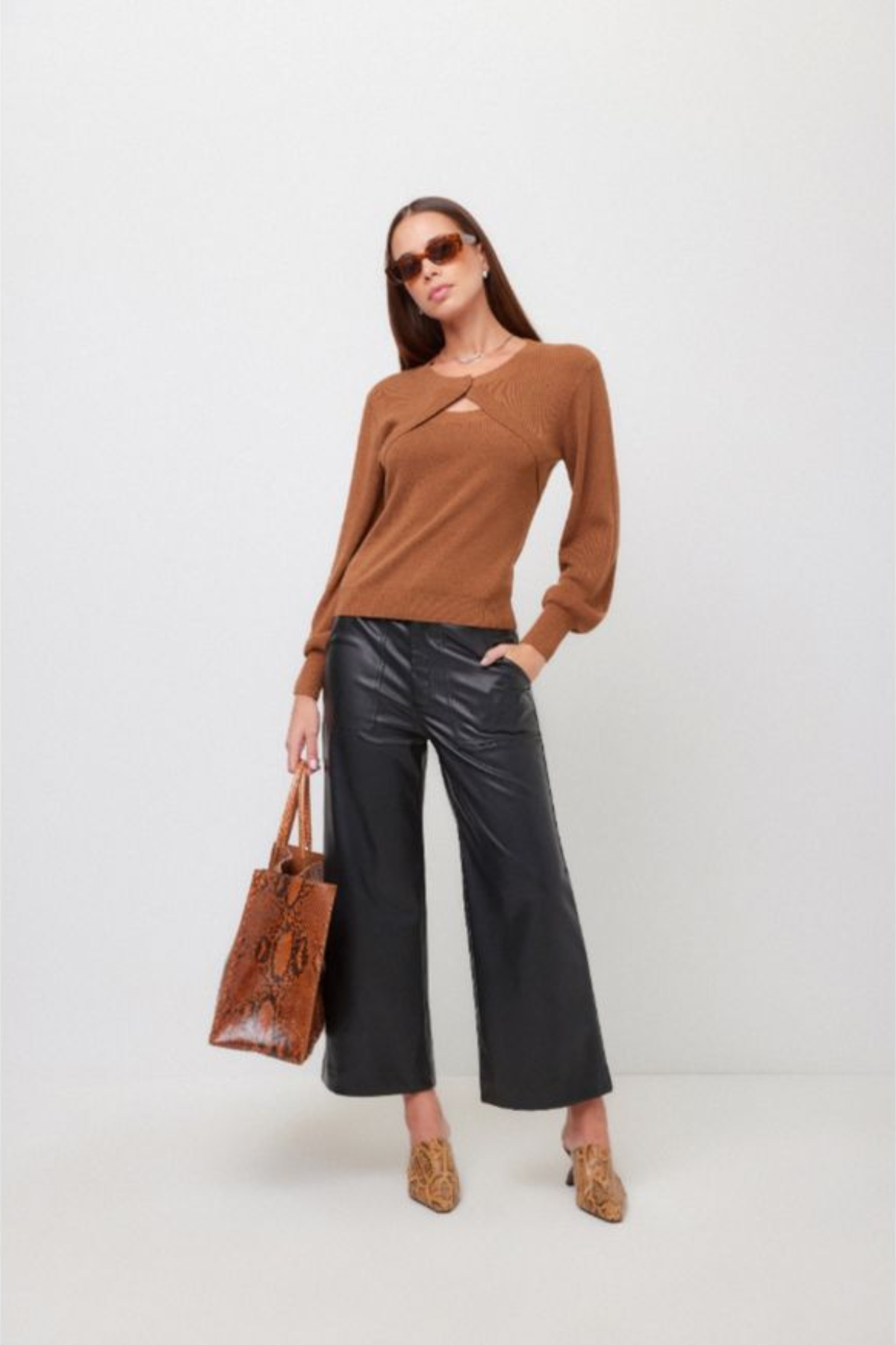 A woman modeling a brown long-sleeve knit top with a crossover front and keyhole cutout, paired with black wide-leg pants, leopard-print heeled mules, sunglasses, and a brown textured tote bag against a plain light gray studio background.