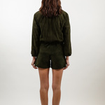 A woman viewed from behind wearing an olive green long-sleeve top and matching shorts set, paired with white heels, against a plain white studio background.