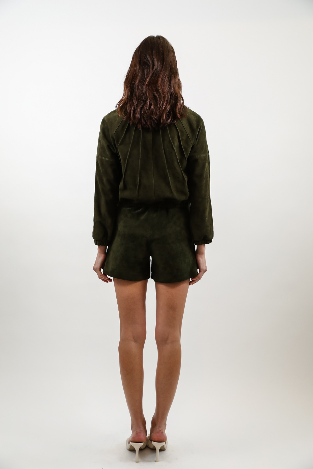 A woman viewed from behind wearing an olive green long-sleeve top and matching shorts set, paired with white heels, against a plain white studio background.