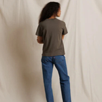 A person standing with their back to the camera, wearing a dark olive green short-sleeve t-shirt and blue jeans, photographed against a draped off-white fabric backdrop.