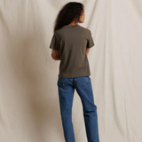 A person standing with their back to the camera, wearing a dark olive green short-sleeve t-shirt and blue jeans, photographed against a draped off-white fabric backdrop.