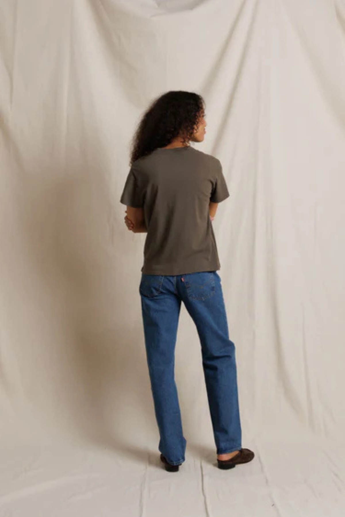 A person standing with their back to the camera, wearing a dark olive green short-sleeve t-shirt and blue jeans, photographed against a draped off-white fabric backdrop.