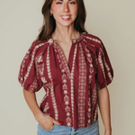 A woman modeling a burgundy short-sleeve blouse with cream vertical floral patterns and a tie-neck detail, paired with light blue jeans against a solid beige background.