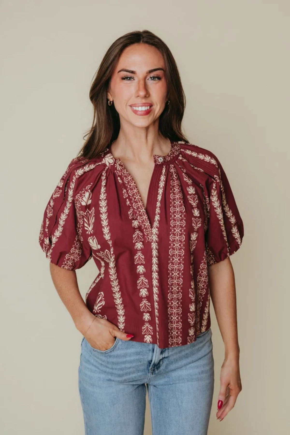 A woman modeling a burgundy short-sleeve blouse with cream vertical floral patterns and a tie-neck detail, paired with light blue jeans against a solid beige background.