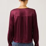 A woman viewed from the back wearing a long-sleeve satin blouse in deep burgundy and blue jeans, set against a plain light gray studio background.