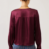A woman viewed from the back wearing a long-sleeve satin blouse in deep burgundy and blue jeans, set against a plain light gray studio background.