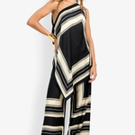 A woman modeling a one-shoulder wide-leg jumpsuit with a black and cream geometric striped pattern, styled with a black beanie, gold bracelets, and black sandals against a plain white studio background.
