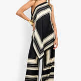 A woman modeling a one-shoulder wide-leg jumpsuit with a black and cream geometric striped pattern, styled with a black beanie, gold bracelets, and black sandals against a plain white studio background.