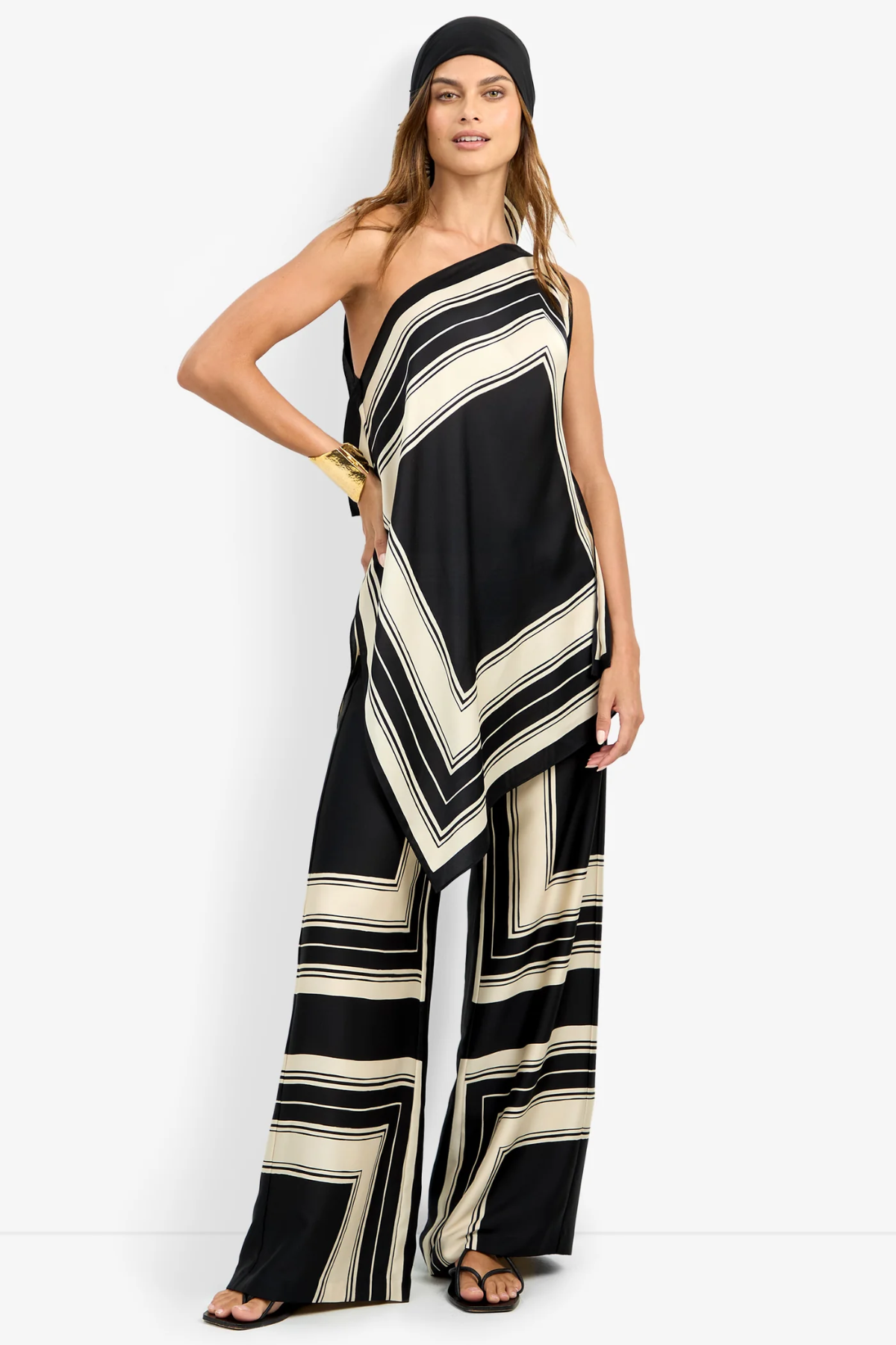 A woman modeling a one-shoulder wide-leg jumpsuit with a black and cream geometric striped pattern, styled with a black beanie, gold bracelets, and black sandals against a plain white studio background.