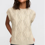 A woman wearing a cream-colored sleeveless knit sweater with a textured geometric pattern, paired with dark blue pants, against a light gray background.