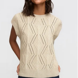 A woman wearing a cream-colored sleeveless knit sweater with a textured geometric pattern, paired with dark blue pants, against a light gray background.