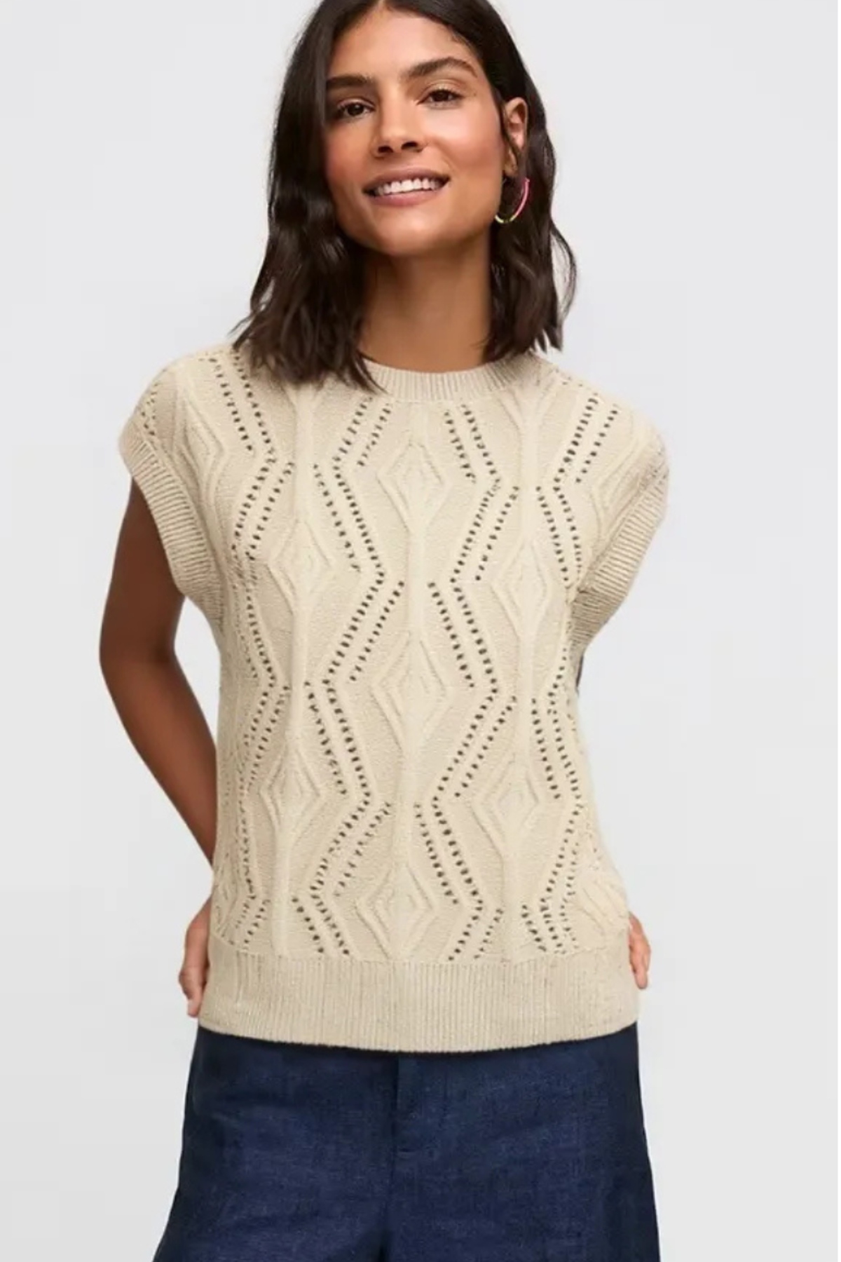 A woman wearing a cream-colored sleeveless knit sweater with a textured geometric pattern, paired with dark blue pants, against a light gray background.