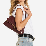 A woman wearing a white sleeveless top and light blue jeans carries a burgundy leather shoulder bag with gold hardware against a plain white background.