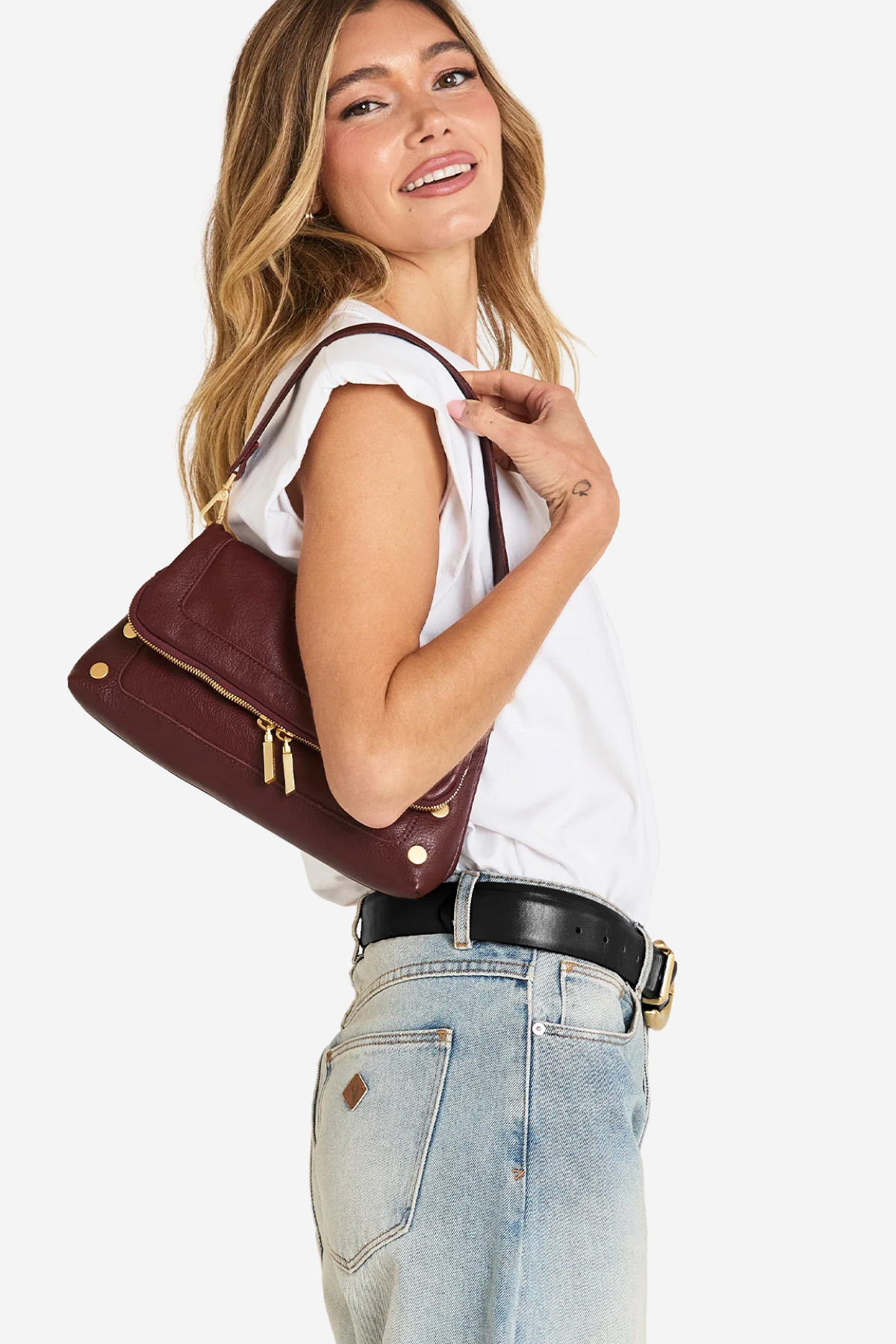 A woman wearing a white sleeveless top and light blue jeans carries a burgundy leather shoulder bag with gold hardware against a plain white background.