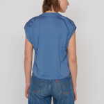 A woman viewed from the back wearing a blue short-sleeve top and blue jeans against a plain light gray studio background.