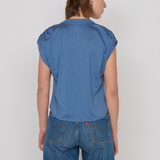 A woman viewed from the back wearing a blue short-sleeve top and blue jeans against a plain light gray studio background.