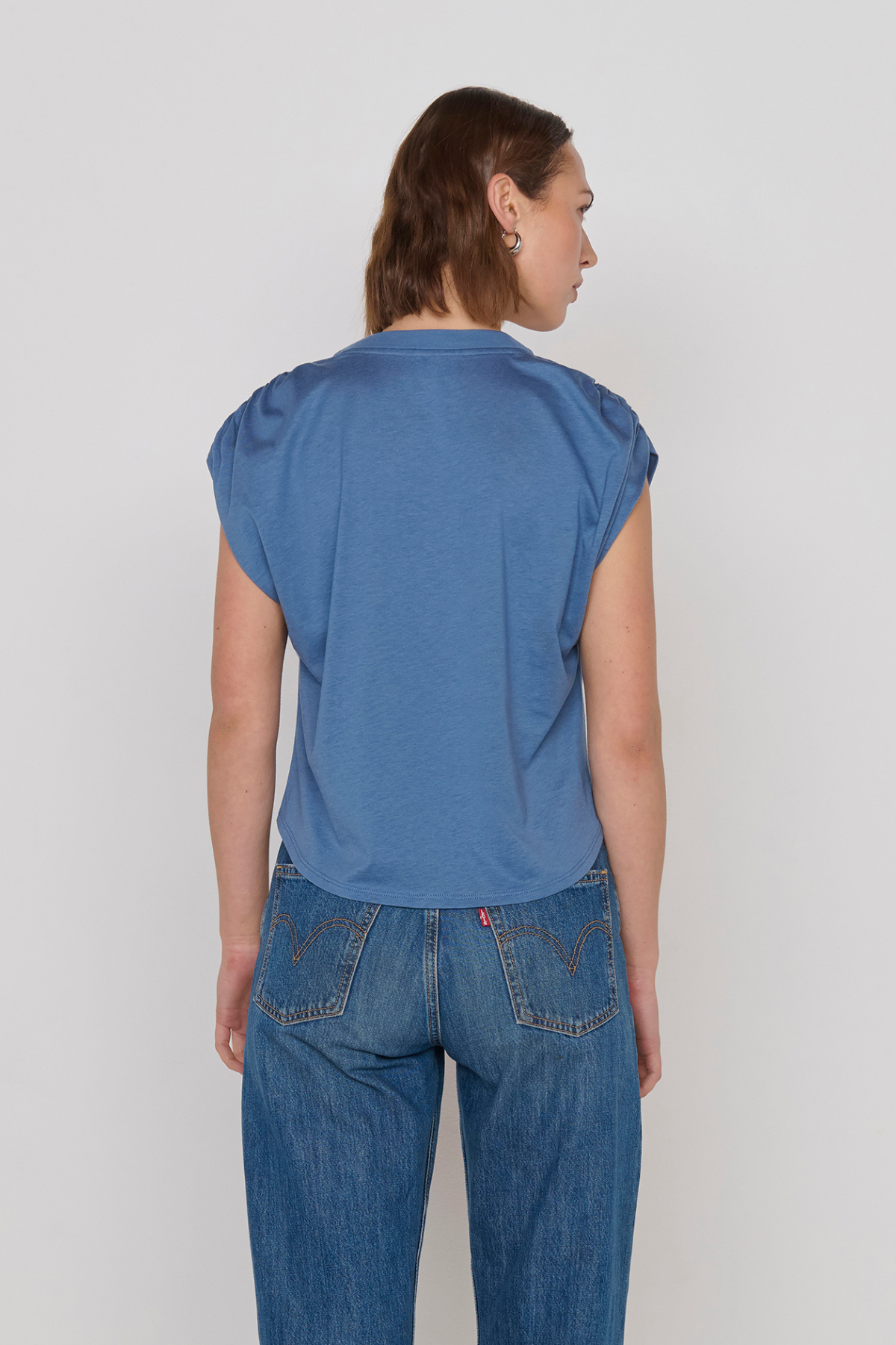 A woman viewed from the back wearing a blue short-sleeve top and blue jeans against a plain light gray studio background.