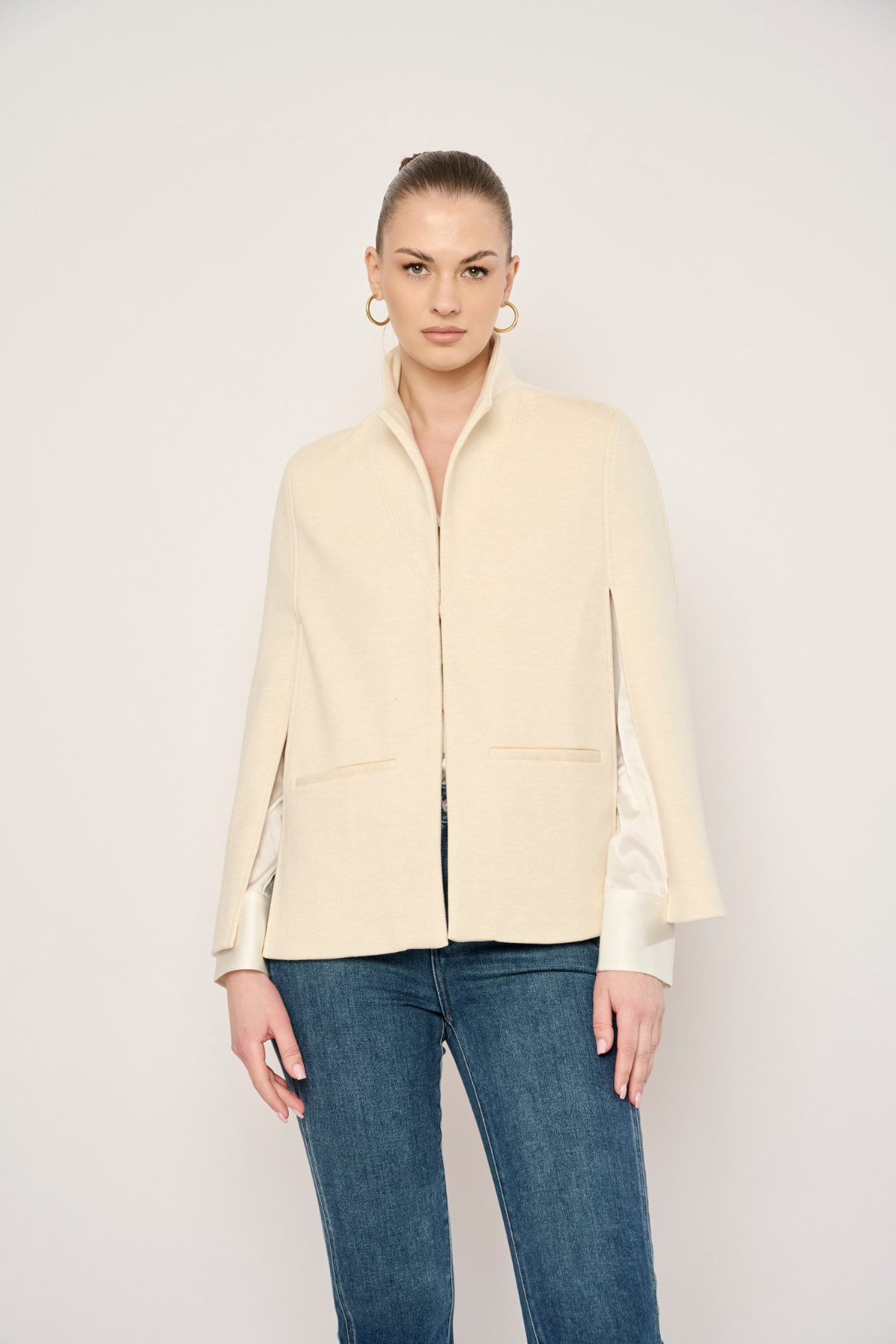 A woman modeling a cream-colored jacket with satin sleeves and blue jeans, standing against a plain light background.