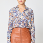 A woman modeling a long-sleeve button-up blouse with a blue, orange, and beige floral paisley pattern, paired with a brown faux-leather skirt, against a light gray studio background.