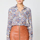 A woman modeling a long-sleeve button-up blouse with a blue, orange, and beige floral paisley pattern, paired with a brown faux-leather skirt, against a light gray studio background.