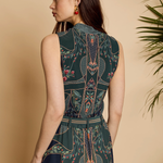 A woman shown from the back wearing a sleeveless dark teal dress with an intricate botanical and geometric print in pink, beige, and white. She is standing against a warm beige wall with a green plant leaf visible on the right side.