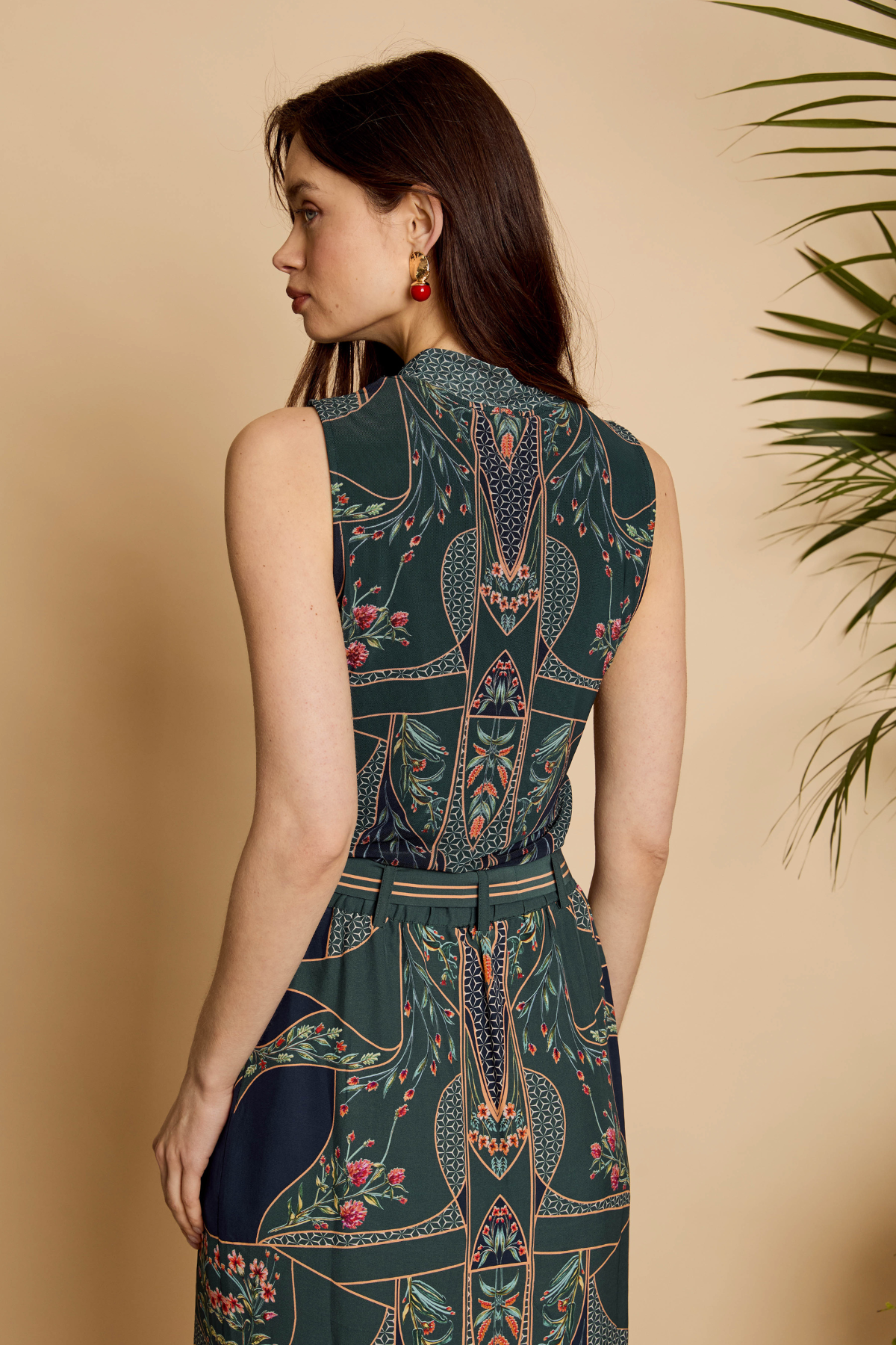 A woman shown from the back wearing a sleeveless dark teal dress with an intricate botanical and geometric print in pink, beige, and white. She is standing against a warm beige wall with a green plant leaf visible on the right side.