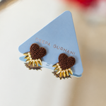 A pair of Deepa Gurnani earrings displayed on a light blue triangular card. The earrings feature a textured dark brown heart-shaped center with a gold-toned sunburst border.