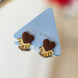 A pair of Deepa Gurnani earrings displayed on a light blue triangular card. The earrings feature a textured dark brown heart-shaped center with a gold-toned sunburst border.