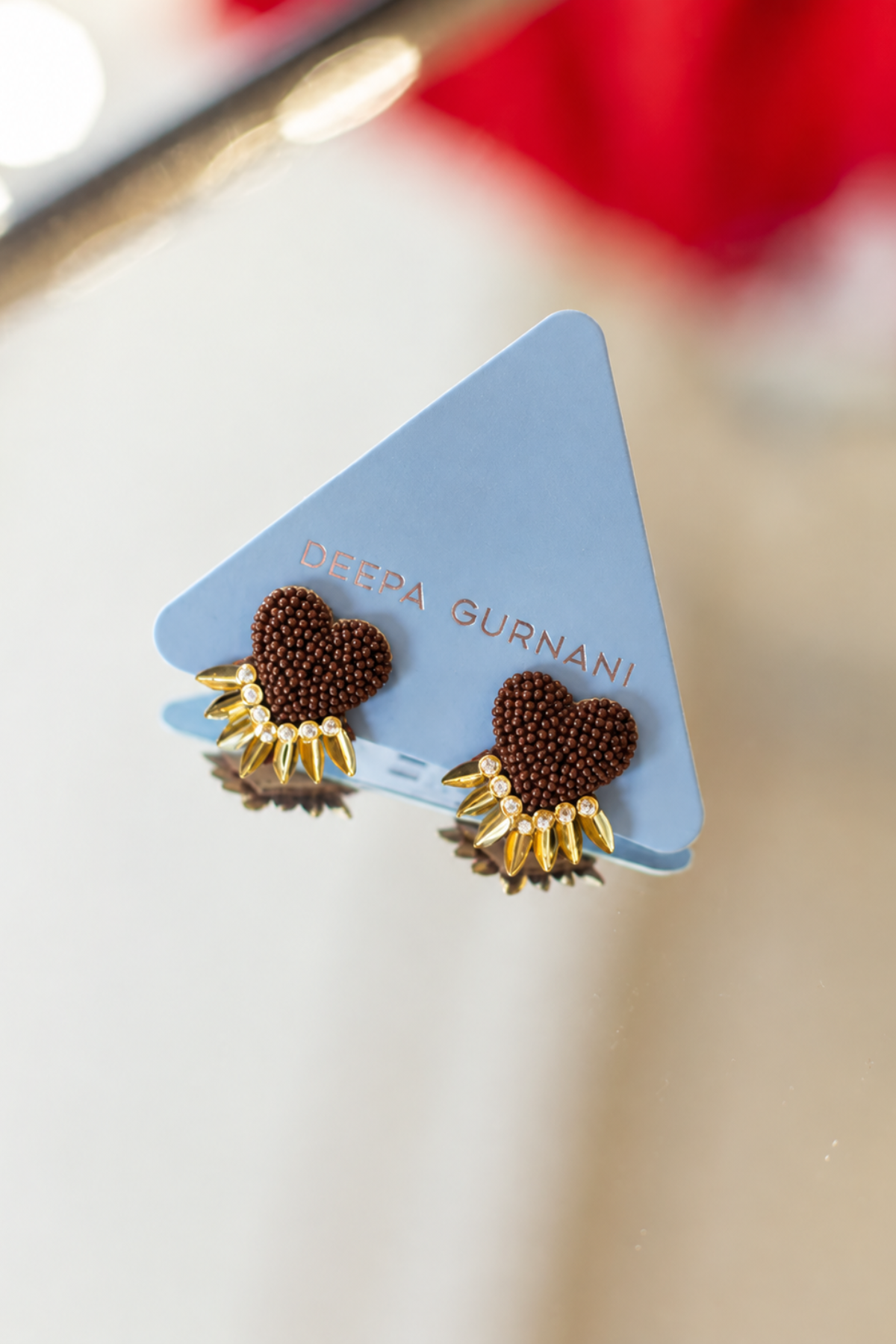 A pair of Deepa Gurnani earrings displayed on a light blue triangular card. The earrings feature a textured dark brown heart-shaped center with a gold-toned sunburst border.