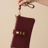 A hand holding a burgundy zip-around pouch with a wrist strap and gold-tone hardware against a solid beige background.