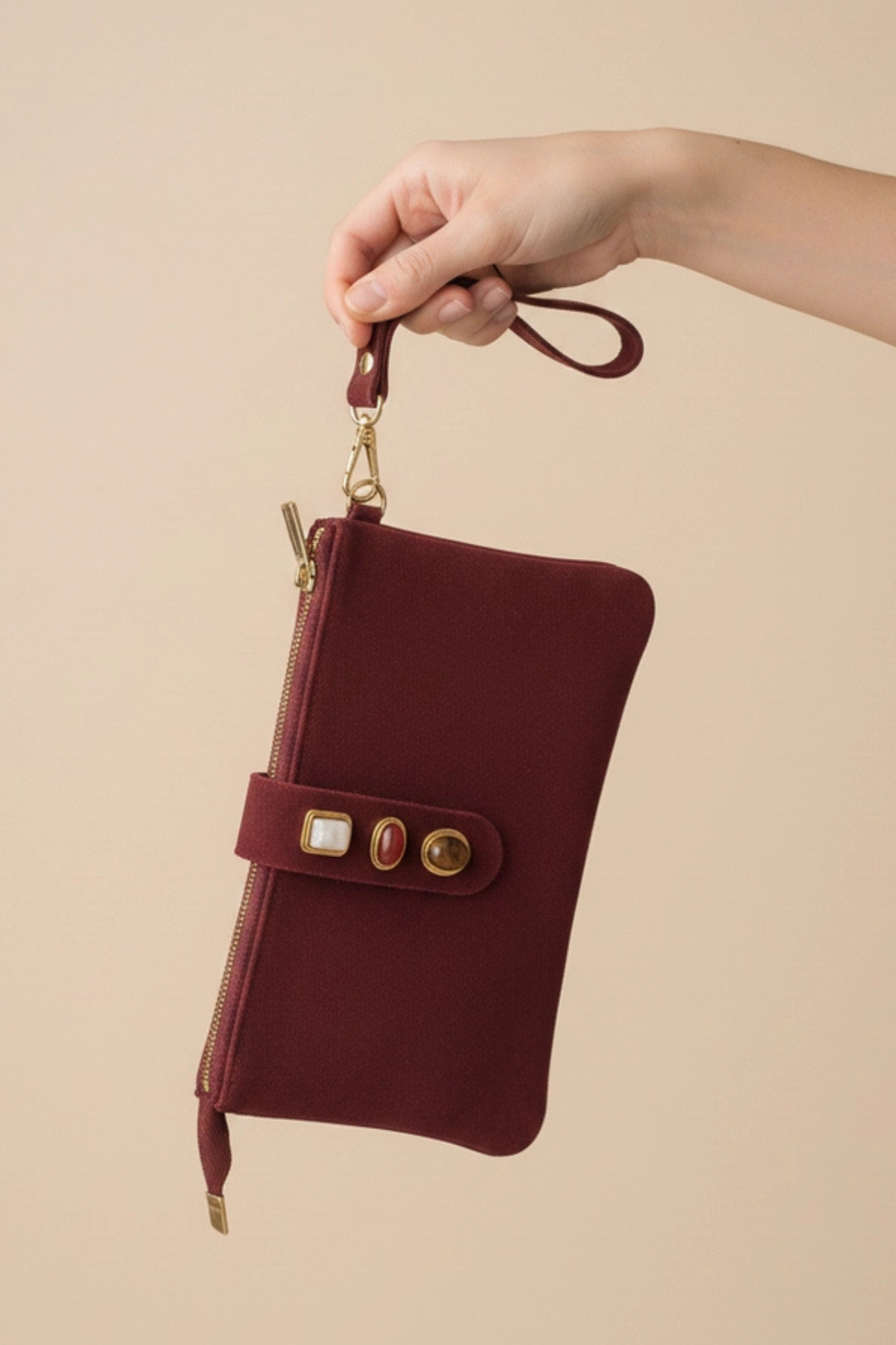 A hand holding a burgundy zip-around pouch with a wrist strap and gold-tone hardware against a solid beige background.
