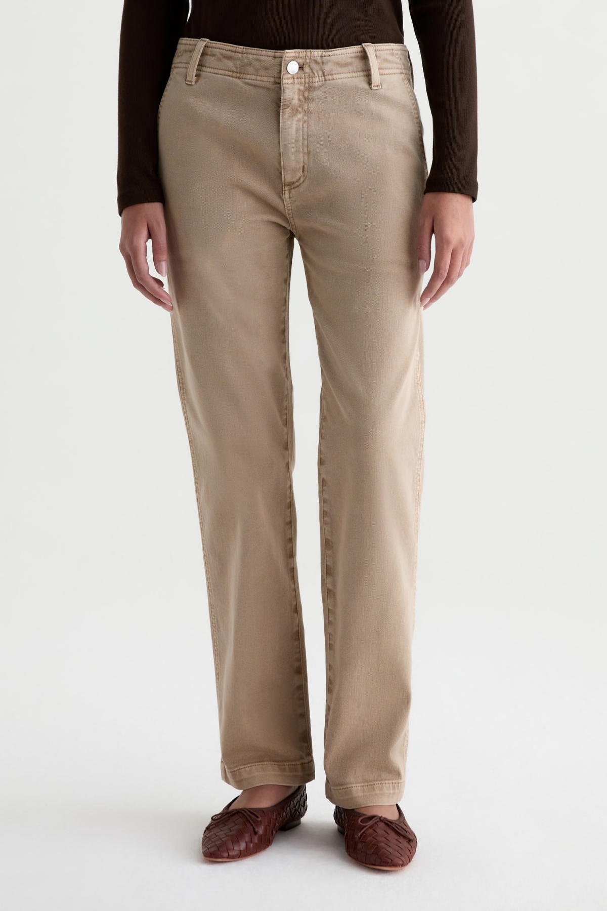A person wearing straight-leg beige jeans with a dark brown long-sleeve top and dark brown woven loafers, shown from the waist down against a light gray studio background.