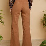 A person shown from the back wearing tan wide-leg pants, a dark embroidered long-sleeve top, and brown heeled sandals, standing against a beige wall with green plants around the scene.