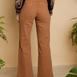 A person shown from the back wearing tan wide-leg pants, a dark embroidered long-sleeve top, and brown heeled sandals, standing against a beige wall with green plants around the scene.