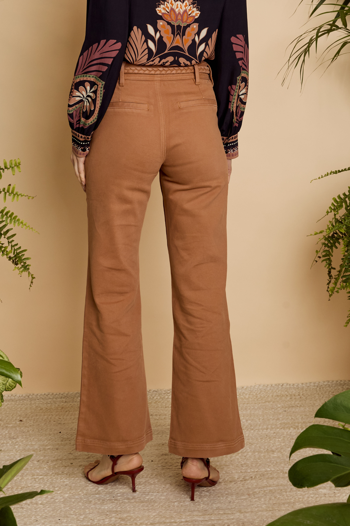 A person shown from the back wearing tan wide-leg pants, a dark embroidered long-sleeve top, and brown heeled sandals, standing against a beige wall with green plants around the scene.