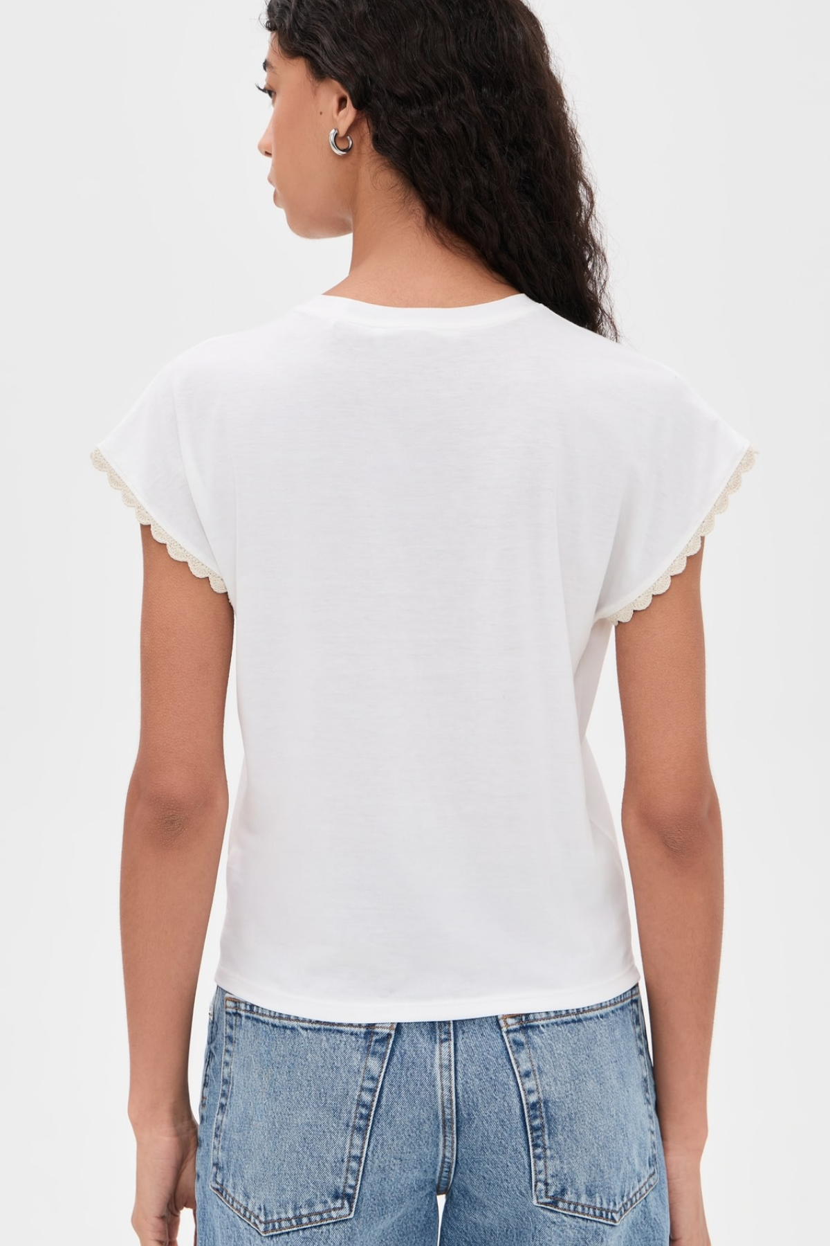 A woman shown from the back wearing a white short-sleeve t-shirt with delicate scalloped edges on the sleeves, paired with light blue jeans, against a plain white background.