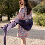 A woman stands outdoors on a gravel path wearing a short, colorful bohemian-style patterned dress and holding a dark purple shawl or wrap.