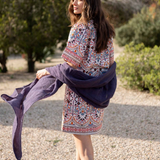 A woman stands outdoors on a gravel path wearing a short, colorful bohemian-style patterned dress and holding a dark purple shawl or wrap.