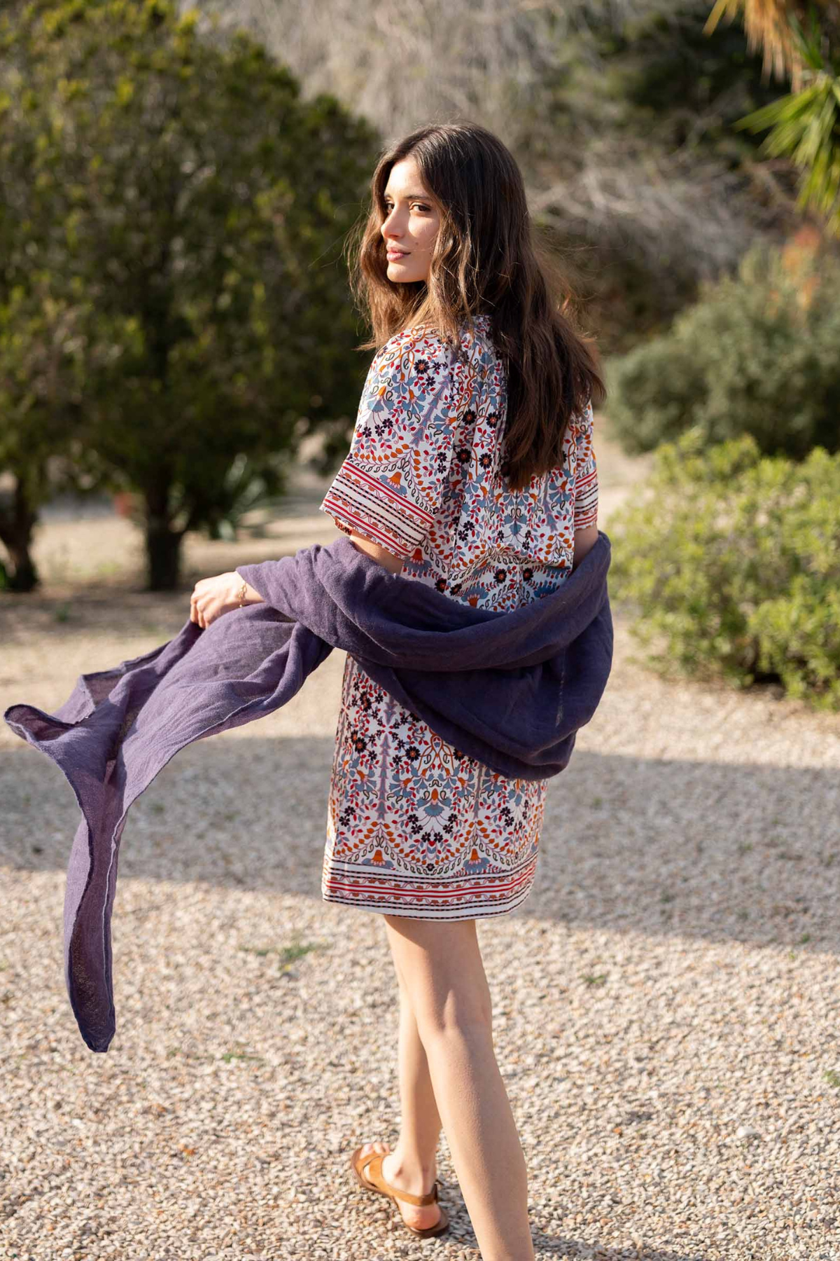 A woman stands outdoors on a gravel path wearing a short, colorful bohemian-style patterned dress and holding a dark purple shawl or wrap.