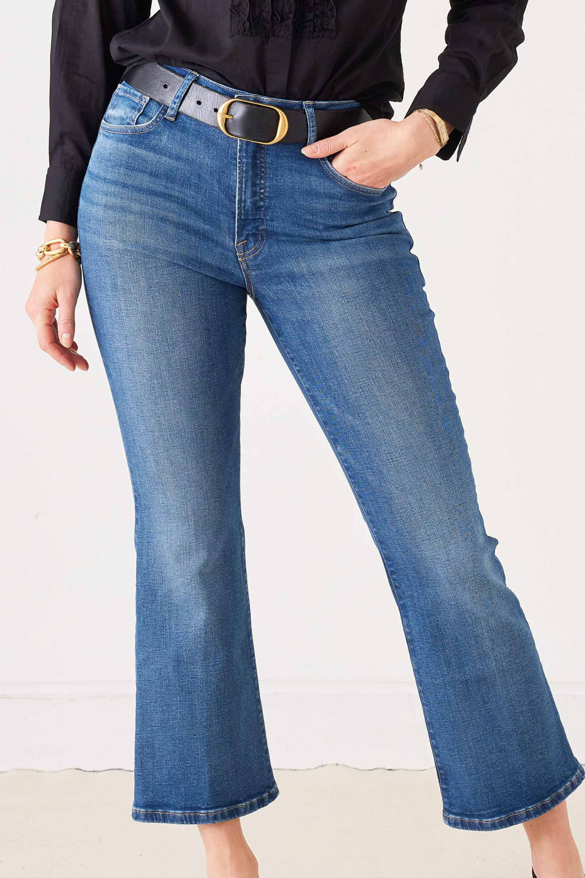 A person wearing medium-wash blue flare jeans with a black belt and gold buckle, paired with a black blouse, shown from the waist down against a plain light background.