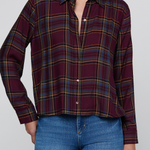 A woman wearing a long-sleeve button-up plaid shirt in deep burgundy with yellow and blue stripes, paired with blue jeans, against a plain light gray background.
