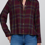 A woman wearing a long-sleeve button-up plaid shirt in deep burgundy with yellow and blue stripes, paired with blue jeans, against a plain light gray background.