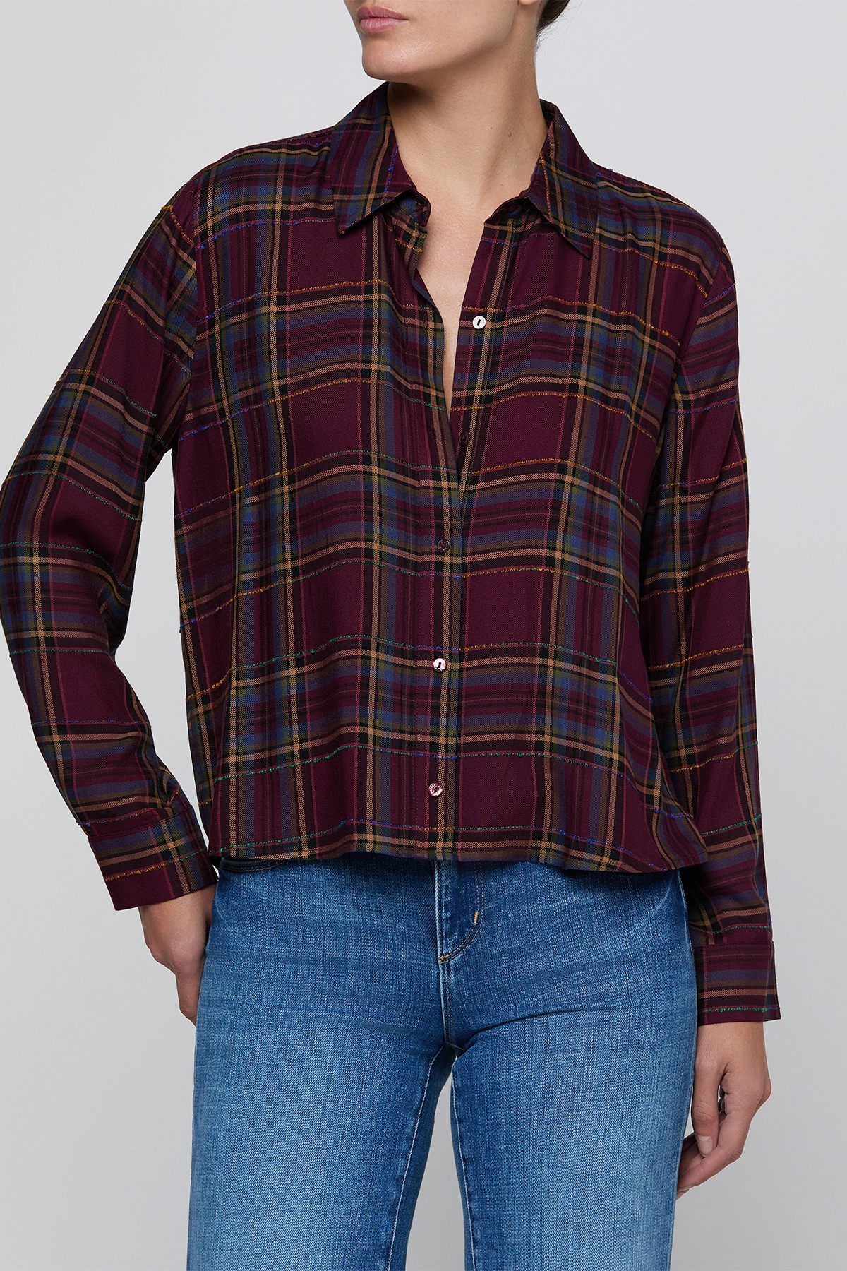 A woman wearing a long-sleeve button-up plaid shirt in deep burgundy with yellow and blue stripes, paired with blue jeans, against a plain light gray background.