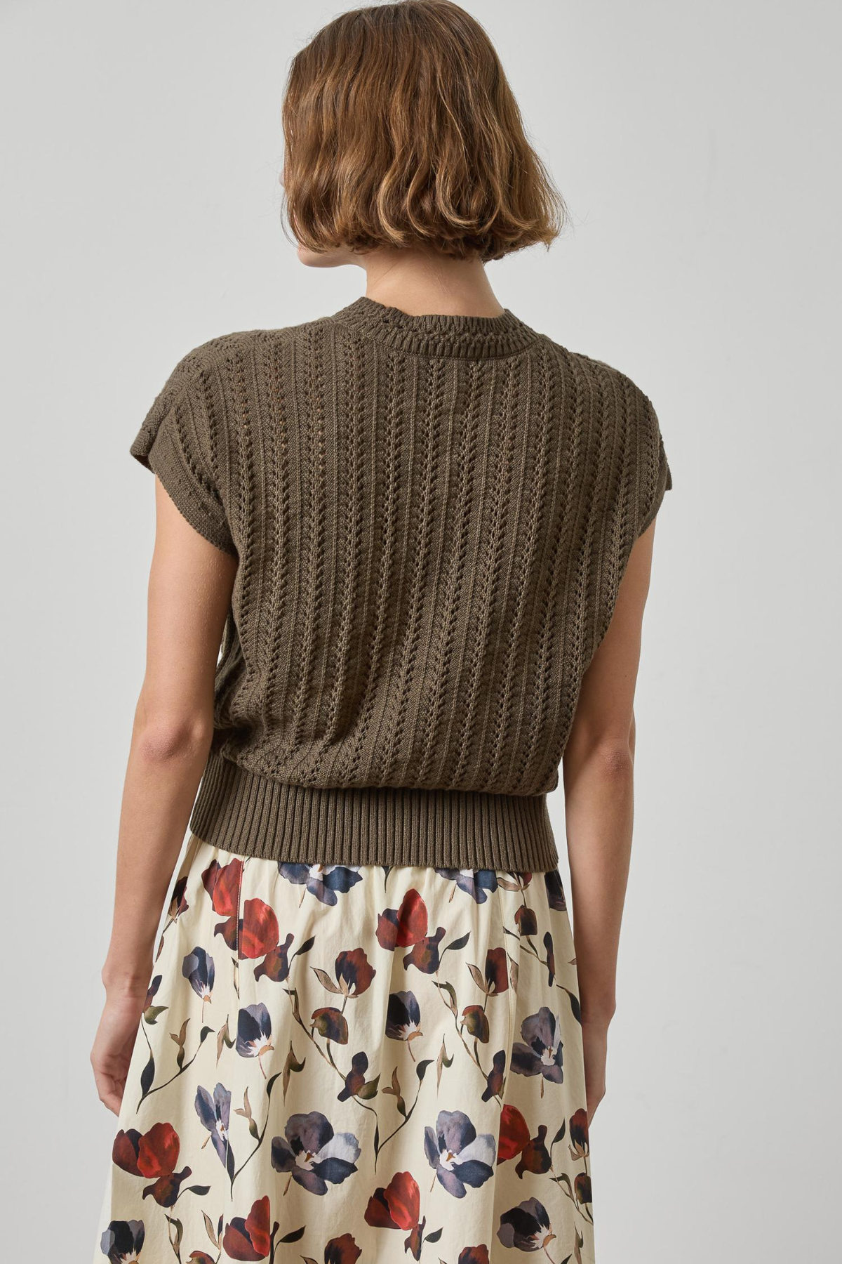 Woman wearing a brown knitted sleeveless sweater over a floral dress on a plain background