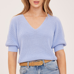 A woman wearing a light blue ribbed knit short-sleeve V-neck top with balloon sleeves, paired with blue jeans and a tan belt, against a plain light beige studio background.