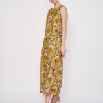 A woman modeling a sleeveless, floor-length dress with a bold yellow, gold, and brown floral pattern against a plain white studio background.